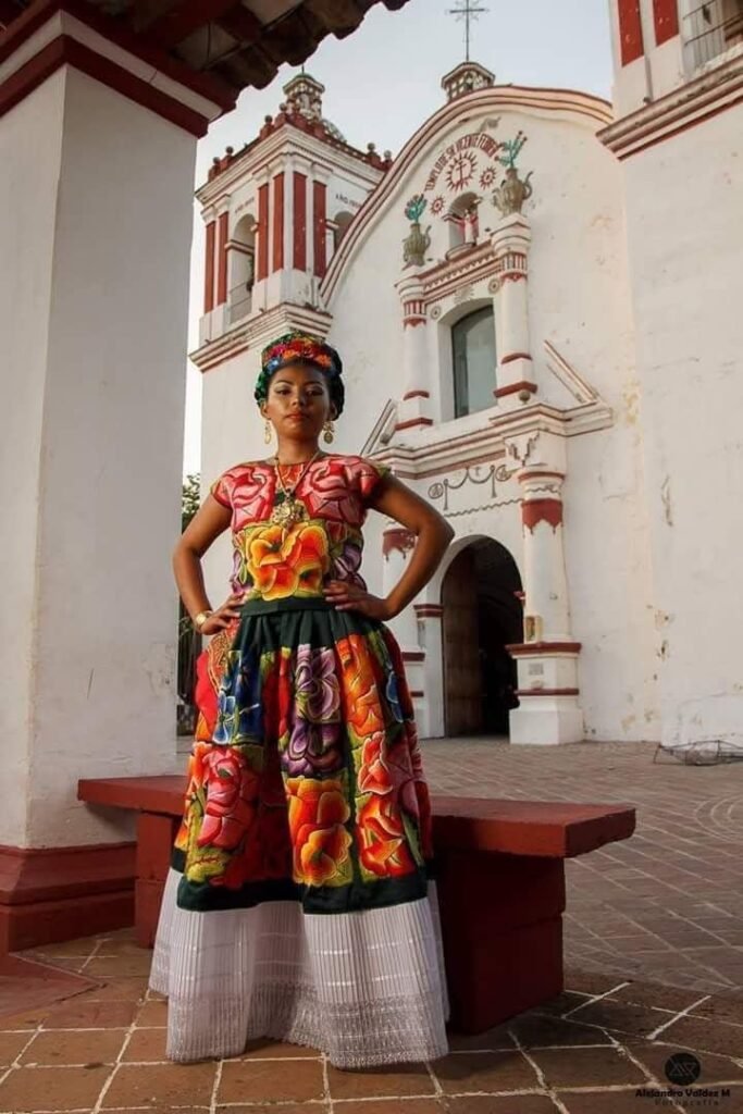 Tehuana en la iglesia Mujer con traje tradicional bordado frente a una iglesia colonial con detalles arquitectónicos